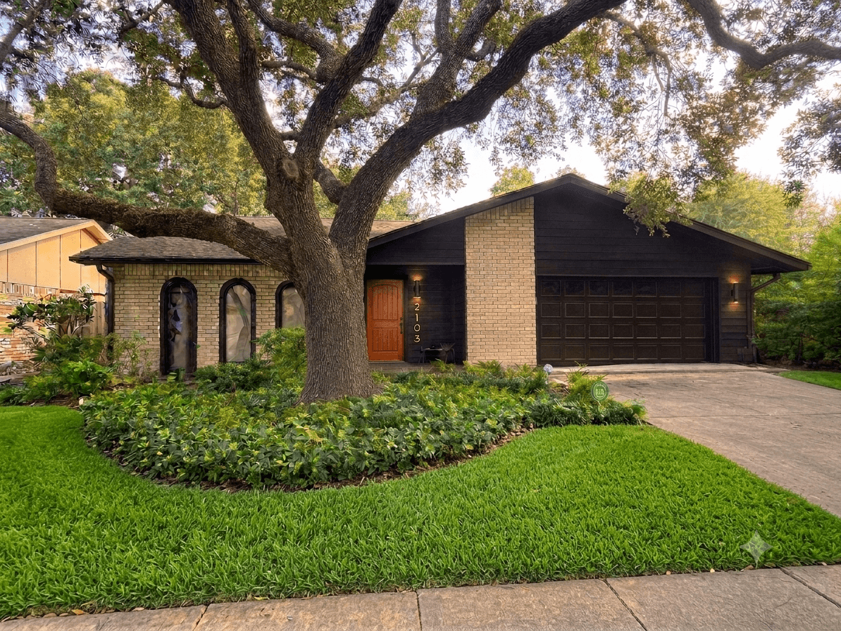 Single-story brick home at 2103 Fall Meadow Dr with dark brown trim, arched front windows, a wood front door, mature live oak shade tree, fresh landscaping beds, and a two-car garage